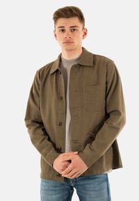 Veste en coton vert olive avec un col, des boutons sur le devant et une poche poitrine, associée à une chemise gris clair et un jean bleu.