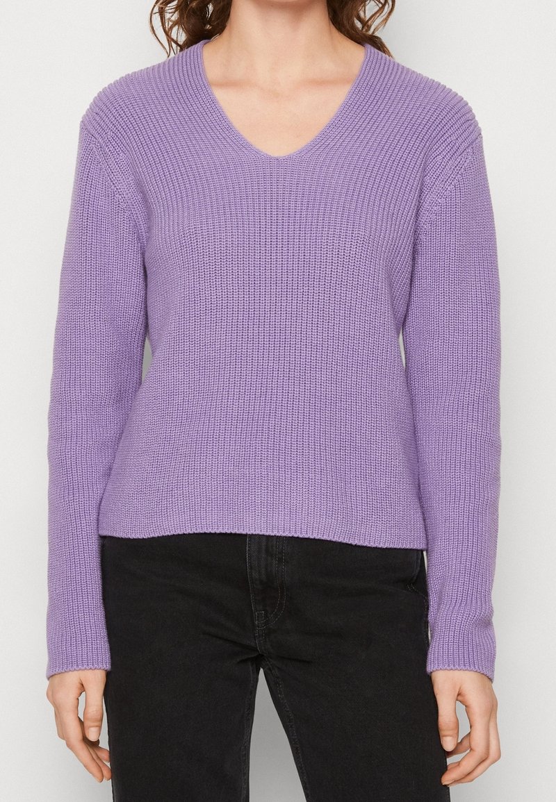 Suéter de punto morado con escote en V, de mangas largas y corte relajado, con un patrón texturizado, combinado con jeans oscuros.