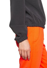 Maglietta grigia a maniche lunghe con polsini elasticizzati abbinata a pantaloni tuta arancioni vivaci dotati di tasche laterali e consistenza liscia.