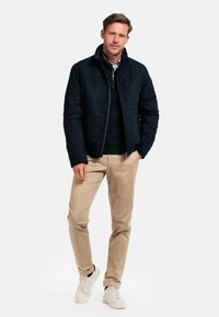 Navy suede jacket with a zip closure, high collar, and side pockets. Paired with beige chinos and white sneakers.