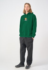 Green hoodie with a front pocket and graphic detail, paired with loose-fitting black pants and black sneakers. Casual design and relaxed fit.