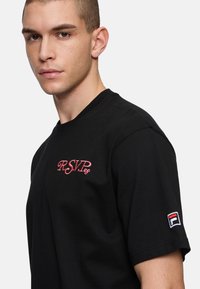 Sort bomulds T-shirt med pink broderet "R.S.V.P." logo på brystet og et Fila logo-patch på venstre ærme. Kortærmet stil.