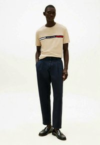 Man wearing beige Tommy Hilfiger t-shirt with logo, navy blue trousers, and black loafers, standing with hands in pockets.