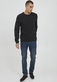 Felpa nera con maniche lunghe, scollo a girocollo e polsini a costine, abbinata a jeans blu slim-fit e scarpe da ginnastica nere.