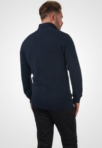 Mørkeblå strikket sweater med høj krave, der har et struktureret ribmønster samt ribkant ved ærmerne og nederst. Vist fra bagsiden.