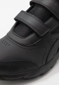 Zapatilla deportiva negra con parte superior de cuero suave, dos correas ajustables de velcro y un panel de tela negra texturizada para mayor transpirabilidad.