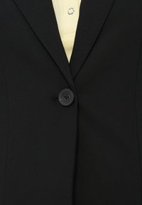 Μαύρο σακάκι με γιακά σε σχήμα V, κλείσιμο με έναν μόνο κουμπί και λεία υφή. Στο εσωτερικό φαίνεται ένα ανοιχτό κίτρινο πουκάμισο από κάτω.