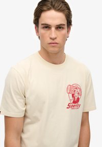 Beige bomuld t-shirt med et rødt grafisk design af en mand med et træstykke. Klassisk rund hals og korte ærmer, blød tekstur.
