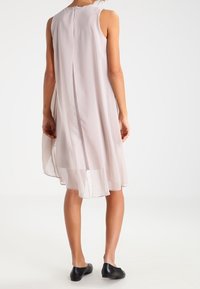 Robe sans manches rose clair avec une silhouette fluide, dotée d'une fente dans le dos et d'un ourlet transparent. Fabriquée en tissu léger, complétée par des chaussures noires.