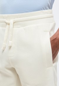 Κοντινό πλάνο ενός ατόμου που φοράει κρεμ υφασμάτινο φούτερ με κορδόνι στη μέση και το χέρι στην δεξιά τσέπη.