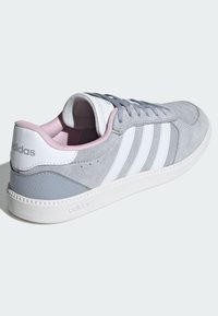 Sivý zamatový a syntetický teniskový topánka s bielymi pruhmi, jemne ružovým vnútrom a textúrovanou podrážkou. Má nízky profil a ploché šnúrky.