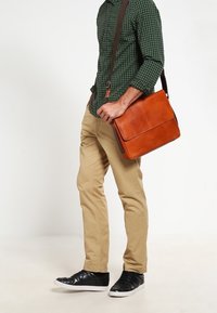 Sac bandoulière en cuir marron avec fermeture à rabat, porté en travers du corps. Modèle portant une chemise à carreaux verte et un pantalon beige, baskets noires.