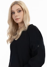 Maglietta nera a maniche lunghe con vestibilità ampia e scollo tondo. Presenta ricami sul petto e un piccolo dettaglio metallico sulla manica.