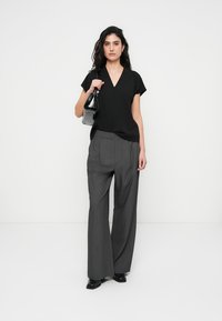 Blouse noire à manches courtes avec un col en V, associée à un pantalon gris taille haute à jambes larges. Le modèle tient un petit sac à main noir.