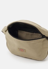 Beige vászonzsák cipzárral, belső zsebével és logóval ellátva. A texturált anyag tartósságot ad.