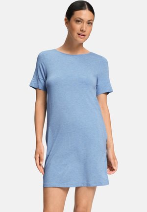 Femme aux cheveux foncés portant une robe bleue claire à manches courtes, debout devant un fond uni clair.