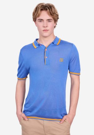 Junger Mann mit blondem Haar, der ein blaues kurzärmeliges Poloshirt mit gelber Einfassung trägt und mit den Händen in den Taschen beiger Hosen steht.