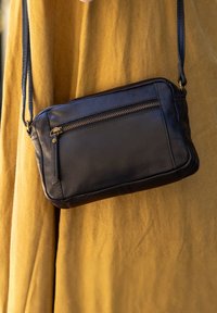 Czarna skórzana torba na ramię z przednią kieszenią na zamek, o płaskim kształcie i smukłym designie. Posiada długi pasek oraz subtelne metaliczne akcenty.