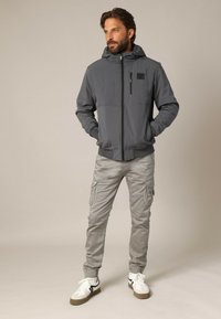 Grijze hooded jack met gewatteerde textuur, voorritssluiting en zak. Gecombineerd met lichtgrijze cargobroek en witte sneakers met zwarte accenten.