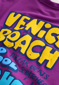 Tissu violet avec un texte en relief jaune et bleu vif indiquant "VENICE BEACH" et un texte bleu plus petit en dessous disant "BREAK BEATS SCRATCHES."