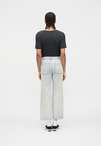 Άτομο με πλεξούδες, που στέκεται με την πλάτη προς τα εμπρός, φοράει μαύρο μπλουζάκι, ανοιχτόχρωμα φαρδιά κοντά τζιν παντελόνια, λευκές κάλτσες και μαύρα αθλητικά παπούτσια.