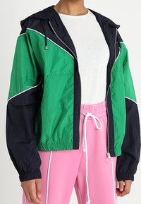 Person iført en grøn og sort zip-up jakke over en hvid ribbet skjorte og lyserøde bukser med snoretræk og hvide piping detaljer.