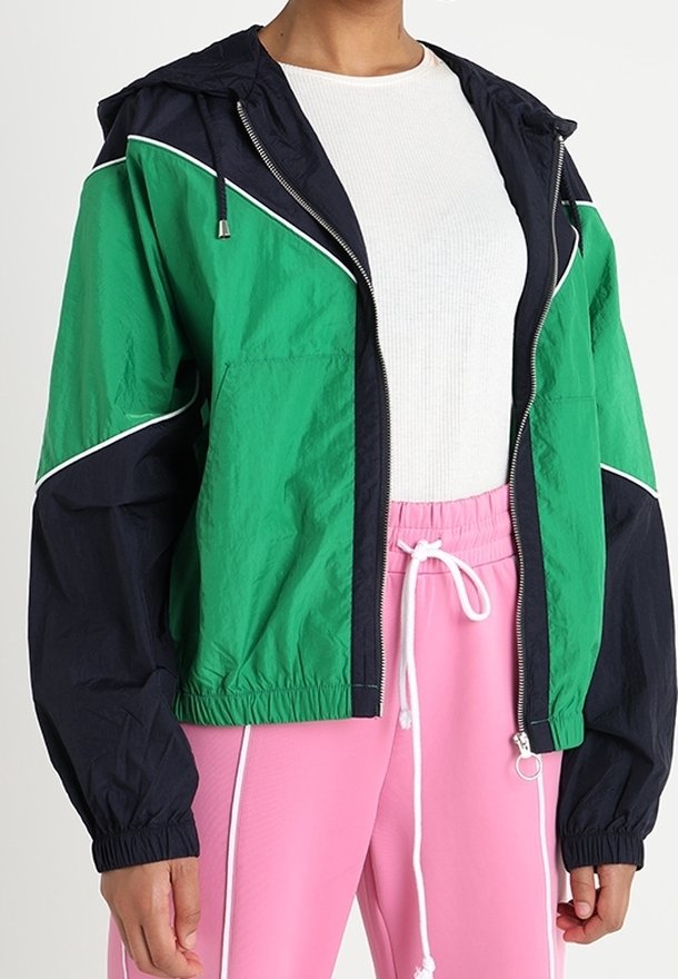 Person iført en grøn og sort zip-up jakke over en hvid ribbet skjorte og lyserøde bukser med snoretræk og hvide piping detaljer.
