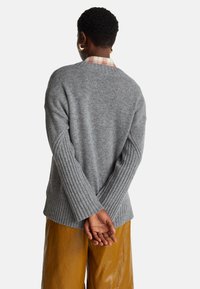 Šedý pletený svetr s žebrovanými manžetami a lemem, doplněný o žluté, lesklé kalhoty s širokými nohavicemi. Pohled zezadu, ruce zkřížené v pase.