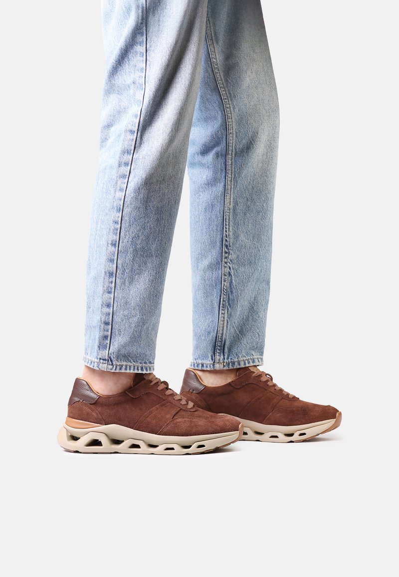 Personne portant un jean bleu clair et des baskets en daim marron avec des semelles beiges, debout devant un fond blanc uni.