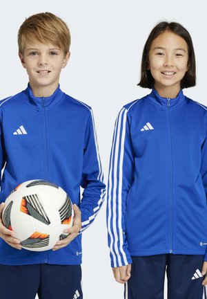 Blaue Sportjacken mit weißen Streifen an den Ärmeln, getragen von zwei Kindern, die einen schwarz-weißen Fußball halten und vor einem einfarbigen Hintergrund stehen.