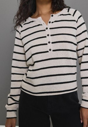 Femme portant une chemise crème à manches longues avec des rayures horizontales noires et des boutons noirs sur le devant, associée à un pantalon noir, sur un fond gris.