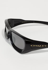 Óculos de sol de desporto Oakley pretos com lentes escuras envolventes e logotipo dourado na ponte, fotografados sobre um fundo branco.