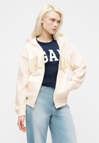 Νέα γυναίκα με μακριά ξανθά μαλλιά, φορώντας κρεμ φούτερ με φερμουάρ πάνω από μπλε μπλουζάκι GAP με λογότυπο και ανοιχτό μπλε τζιν, ποζάροντας μπροστά σε λευκό φόντο.