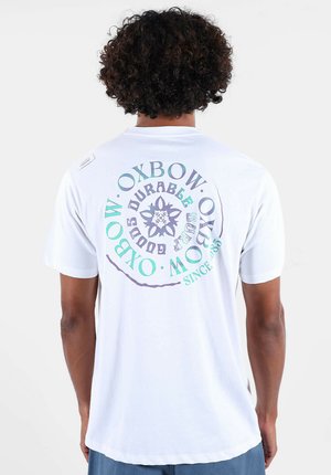 T-shirt en coton blanc avec un motif circulaire au dos en violet et turquoise, accompagné de texte et d'éléments décoratifs.