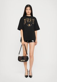 Black oversized t-shirt with gold metallic "JUST CAVALLI" print, paired with shiny black shorts and a textured black handbag. Black heels.
