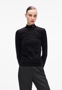 Donna con capelli lisci e indietro che indossa un top lungo a maniche nere scintillante a collo alto e pantaloni neri, su uno sfondo bianco semplice.