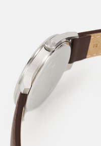 Cadran de montre en métal argenté avec une finition brossée, doté d'une couronne striée et d'un bracelet en cuir brun foncé avec des coutures claires.