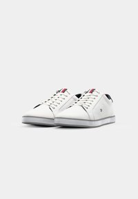 Ett par vita låga sneakers med röda och blå ränder på tungan, snörning och en liten sidologotyp, mot en enkel ljus bakgrund.