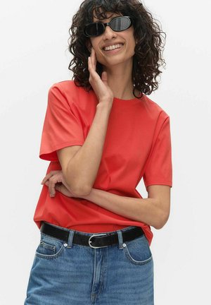 Femme aux cheveux bouclés portant des lunettes de soleil ovales noires, un t-shirt rouge à manches courtes, un jean bleu et une ceinture noire, souriante et touchant son visage.