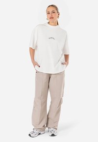 Hvit oversized t-skjorte med ordet "Pegador" brodert i grått, sammen med beige løse bukser og hvite joggesko med svarte detaljer.