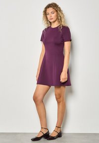 Lila Kleid mit kurzen Ärmeln, tailliertem Oberteil und schwingendem Rock. Hergestellt aus glattem, elastischem Material. Ein Paar schwarze Schuhe mit Riemen am Knöchel.