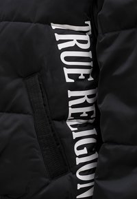 Czarna kurtka puchowa z gładką fakturą, pikowanym designem i wyraźnym białym logo z boku. Posiada boczną kieszeń z klapką.