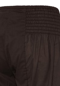 Gros plan sur un pantalon en tissu brun foncé avec une taille élastiquée à smocks et un tissu ample et froncé en dessous de la taille.