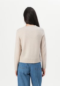Beige, strikket genser med rund hals og lange ermer. Stoffet ser glatt ut med en subtil tekstur, og slutter over høy midje, blå jeans.