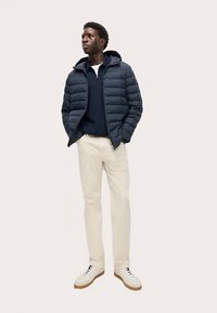 Marineblå dunjakke med hætte, lagdelt over en mørkeblå skjorte, kombineret med beige bukser og hvide sneakers med brune såler.