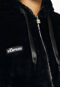 Sort faux fur hættejakke med sølvfarvet lynlås, satin snoretræk og et hvidt broderet "ellesse" logo på forsiden.