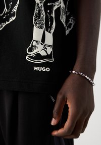 Dark-skinned hand wearing red, white, and silver beaded bracelet beside black clothing with white graphic of sneakers and "HUGO" text.