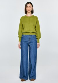 Žena v olivově zeleném zapínaném svetru, širokých modrých džínách a hnědých botách stojí před jednoduchým světlým pozadím.