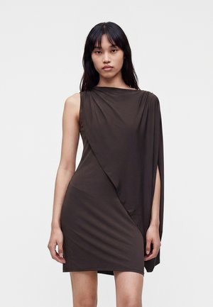Femme aux longs cheveux noirs portant une robe asymétrique sans manches de couleur marron foncé avec une épaule drapée.