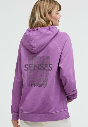 Frau mit blondem Haar, die einen violetten Kapuzenpullover mit der Aufschrift "SENSES" und einem gemusterten Design auf dem Rücken trägt, seitlich zugewandt.
